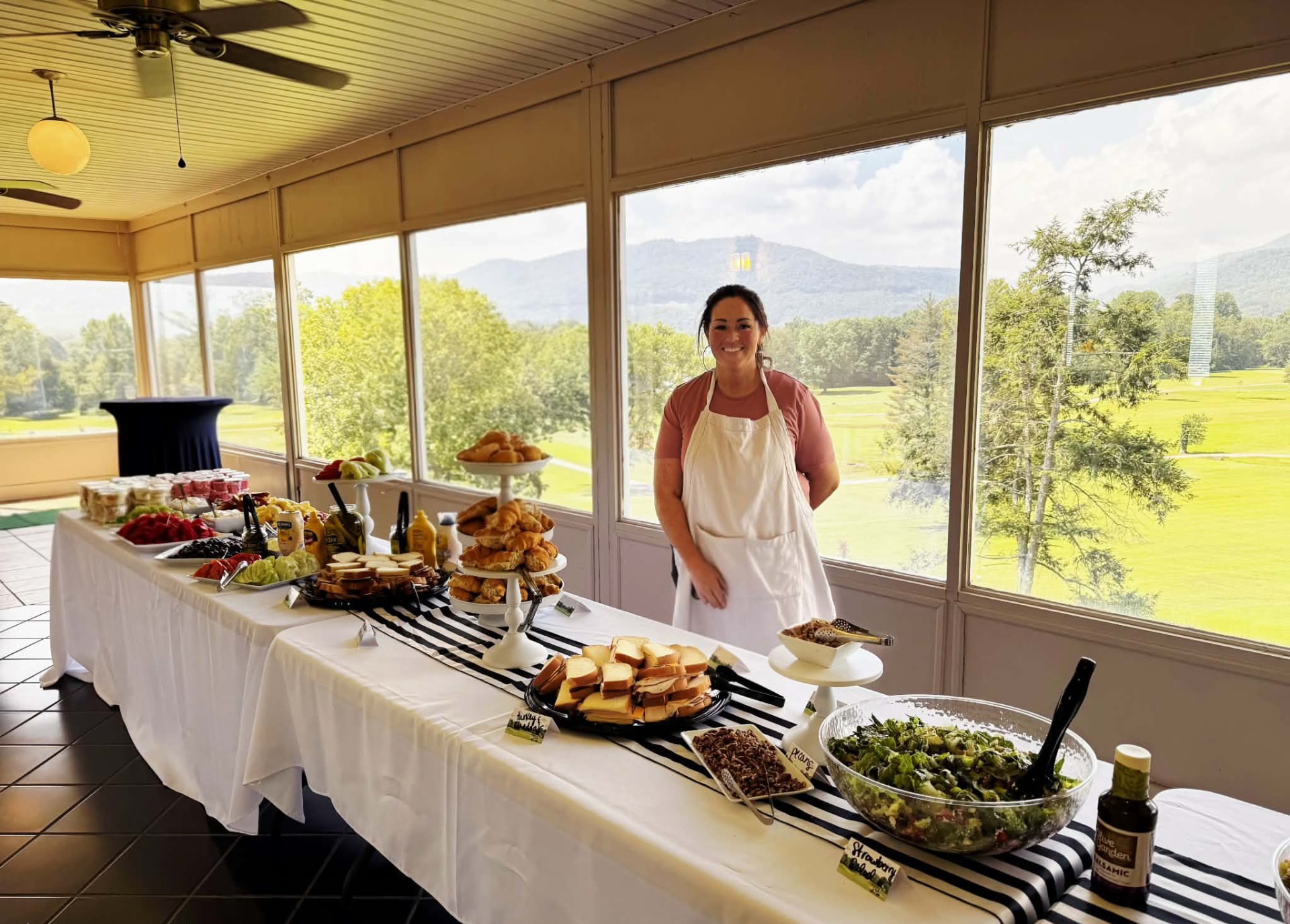 Jeannette of Cater It standing behind a long buffet table of croissants, sandwiches, fresh fruit and salad